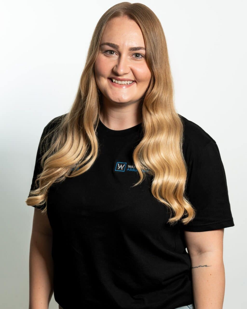 A person with long, wavy blonde hair smiles, wearing a black t-shirt with a small logo.