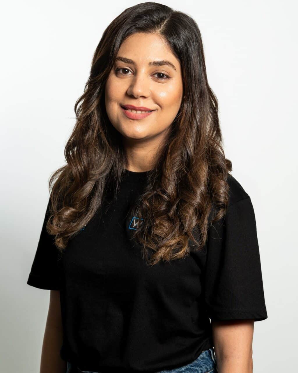 A person with long, wavy hair wearing a black t-shirt is standing against a plain white background.