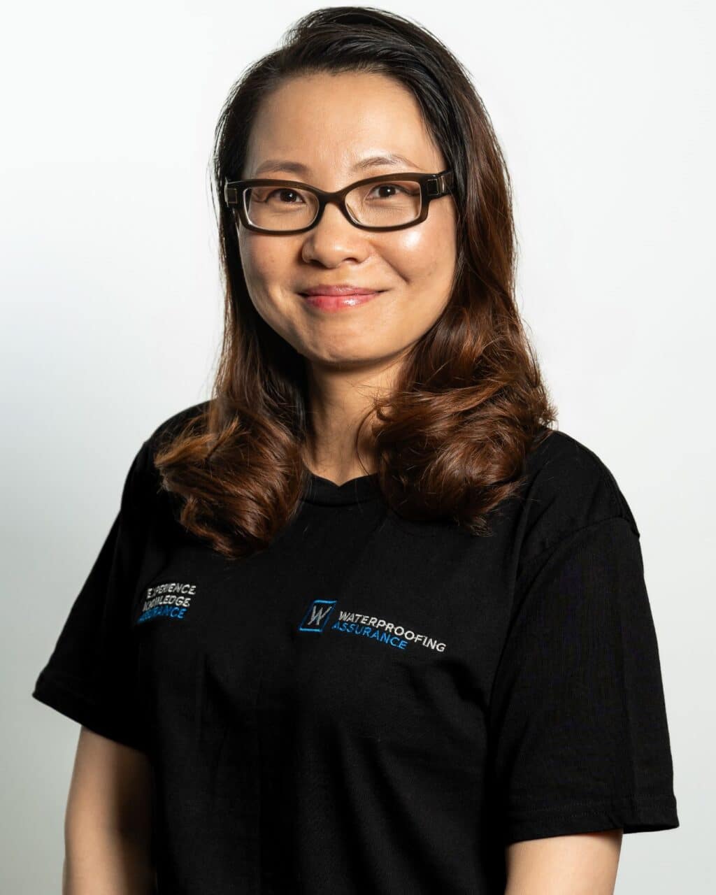 A person with glasses and wavy hair is wearing a black T-shirt with logos related to waterproofing assurance.