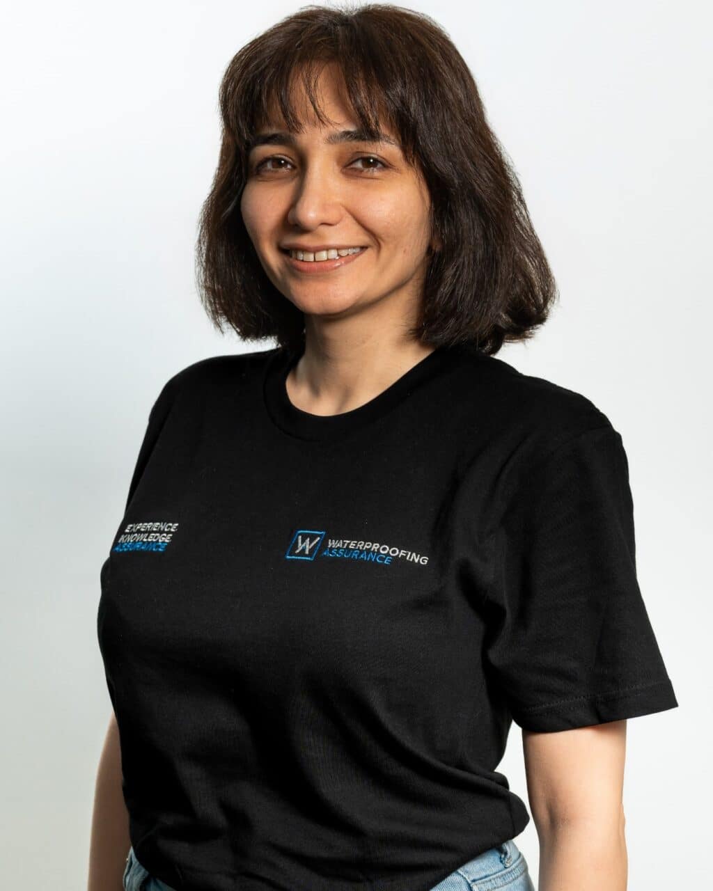 Person with shoulder-length hair wearing a black t-shirt with logos, standing against a light background.