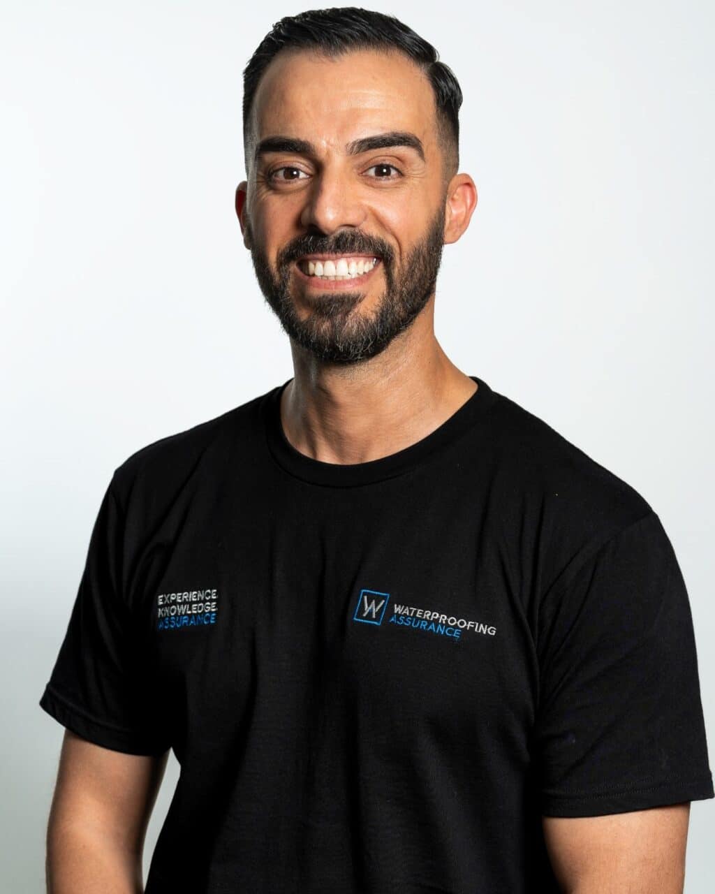 A smiling man with a beard wearing a black T-shirt with the logo and text for "Waterproofing Assurance.