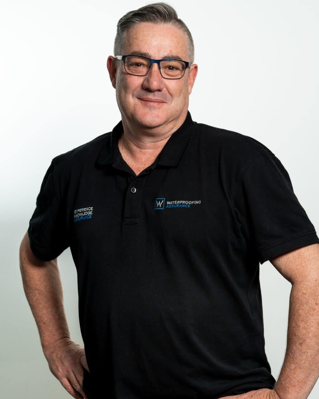 A man wearing glasses and a black polo shirt with embroidered logos poses confidently against a plain background.