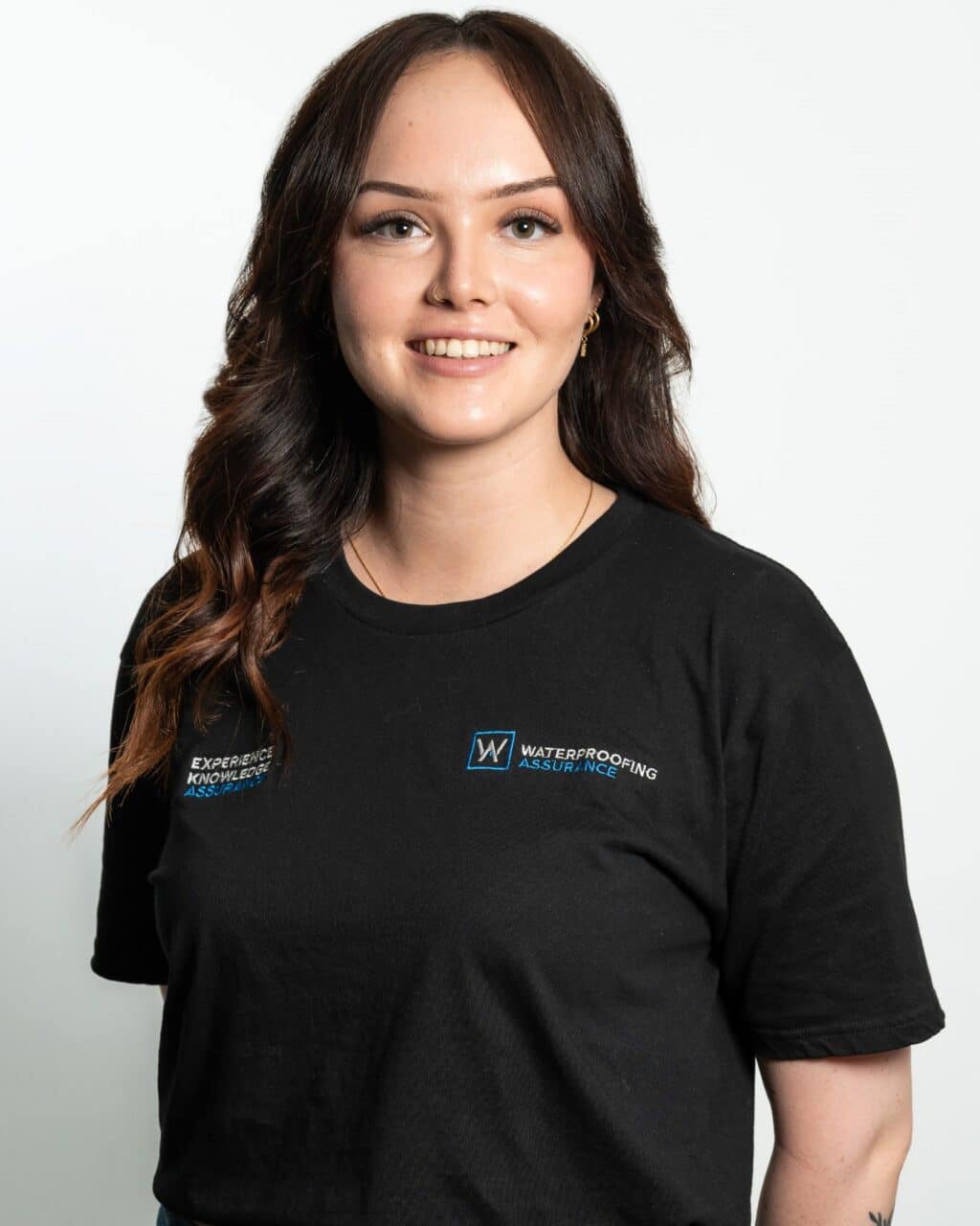 A person with long brown hair is wearing a black T-shirt with "Waterproofing Assurance" printed on it, standing against a plain white background.