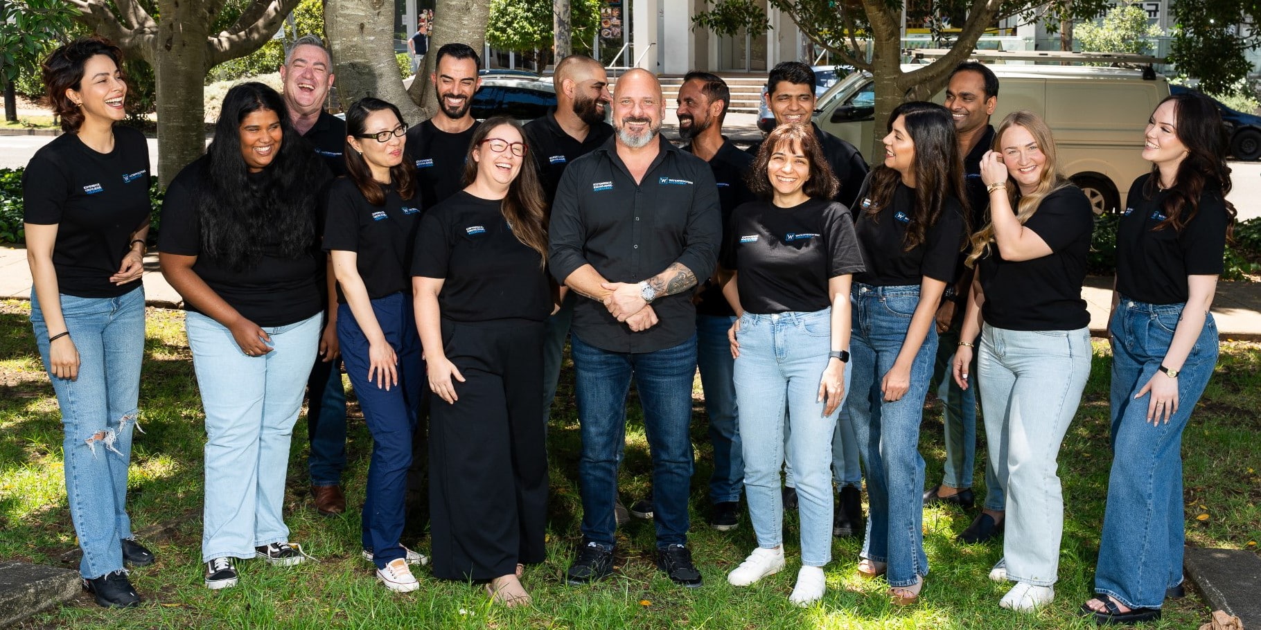 A group of people in black shirts and jeans stand together outdoors, smiling and laughing.