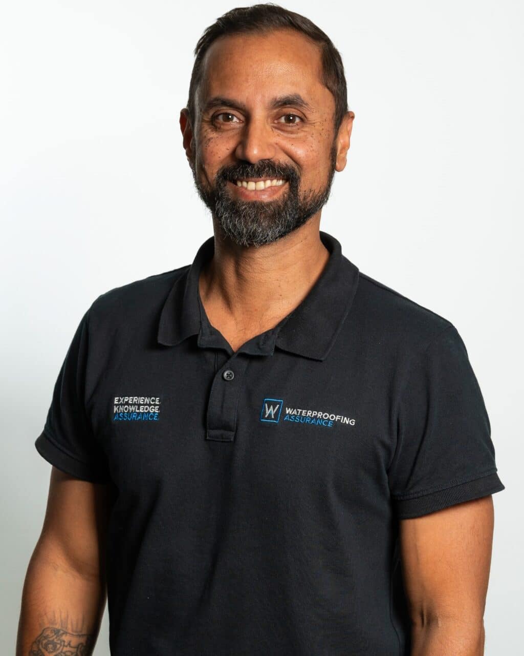 Man in a black polo shirt with company logos, smiling, against a plain white background.