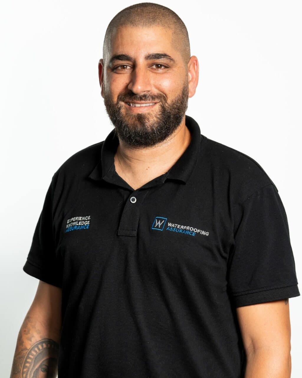 Man with a beard smiling, wearing a black polo shirt with a company logo and tattoo on his arm, standing against a plain background.