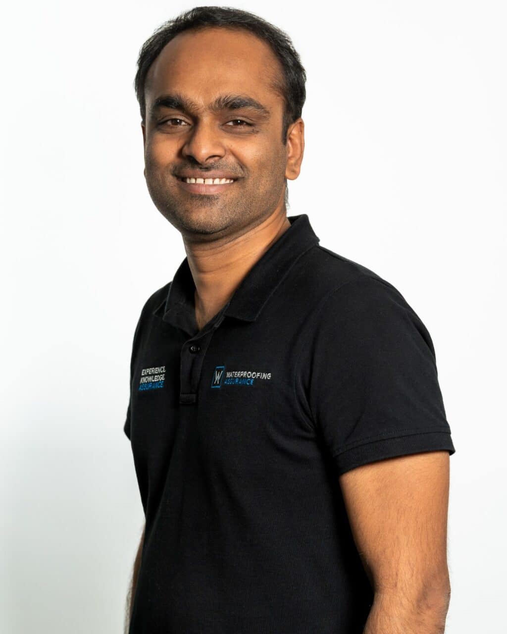 A man in a black polo shirt smiling with arms crossed, standing against a white background.