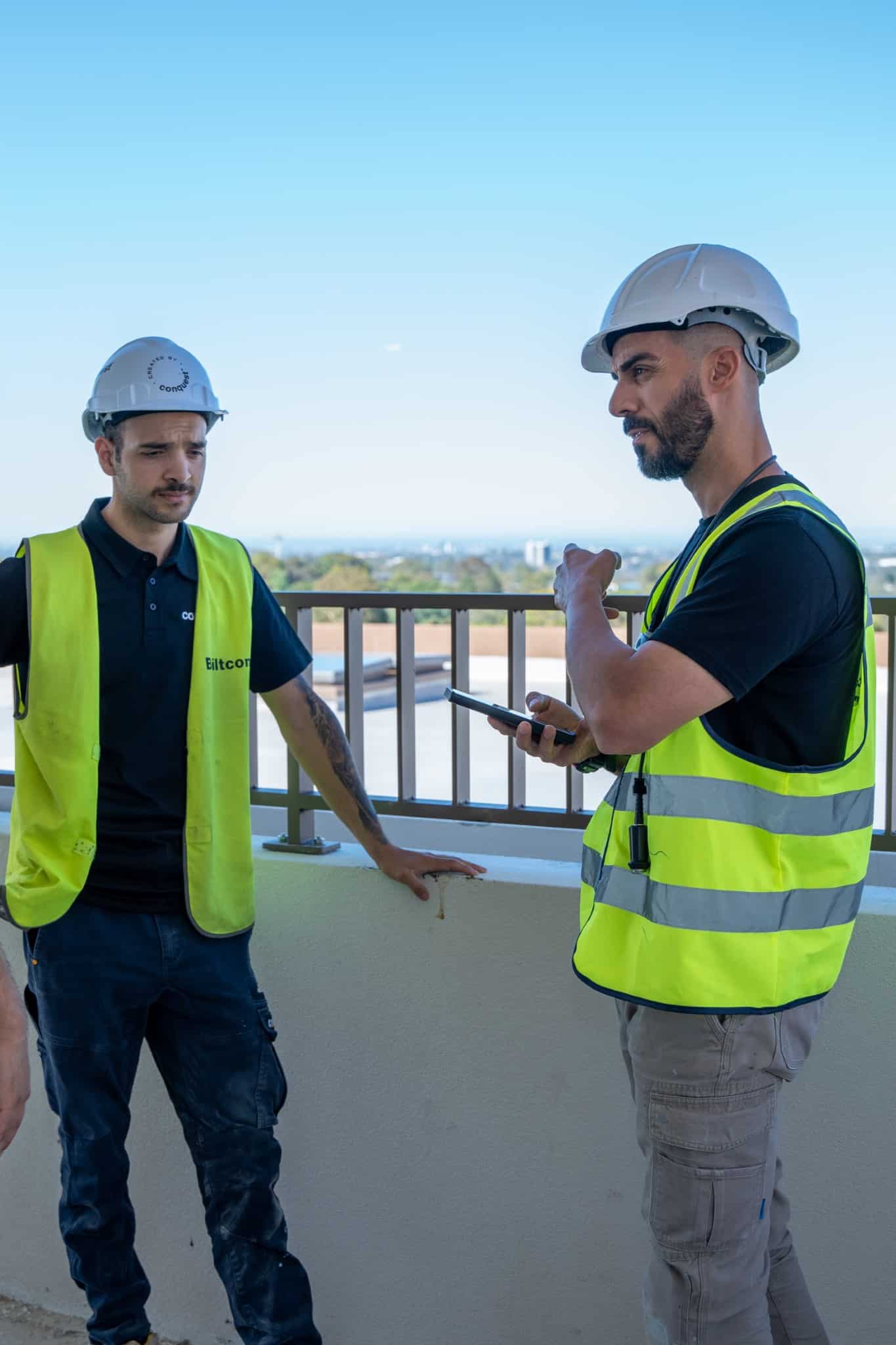 Two construction workers on a rooftop, wearing safety vests and helmets. One holds a smartphone, gesturing while speaking to the other, who rests his hand on a ledge.