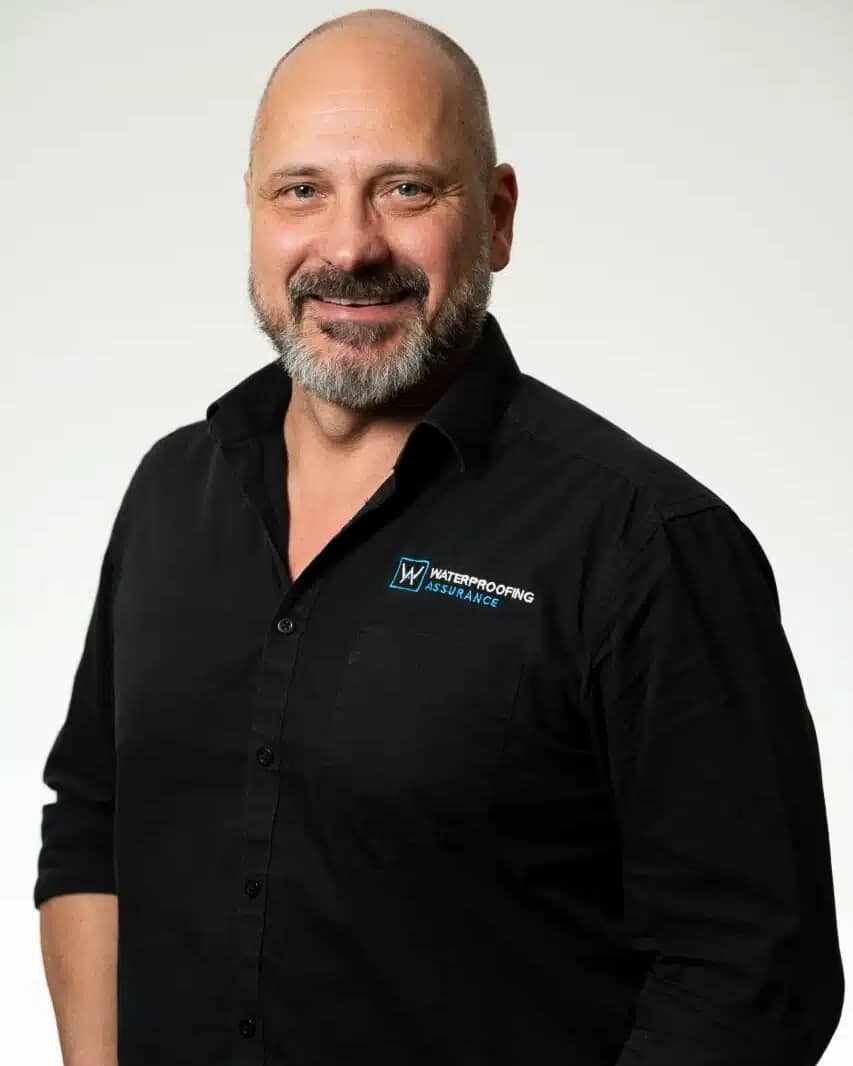 A man with a beard smiles, wearing a black shirt with a company logo on the chest, against a plain background.